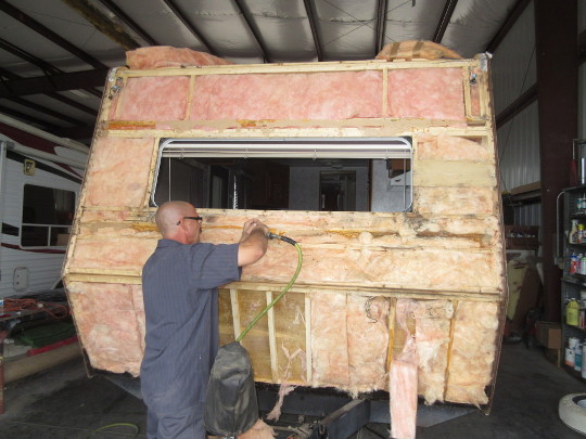 Person aims a hose nozzle at the rear of an RV which is tinted orange, yellow, and red with dirt.